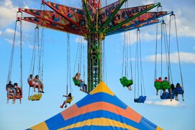 Image of a flying carousel of all different colors. There are people sitting in the chairs with the sky in the background. A blue and orange tent top is emerging from the bottom of the image.