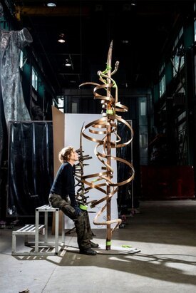 Image of woman sitting and looking at a sculpture of a tall bronze piece with twisting swirls from top to bottom