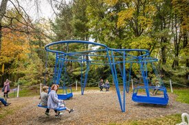 A large, blue abstract cloud-shaped sculpture with three traditional swings and two accessible swings set amongst trees.