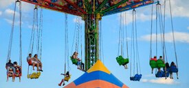 Image of a flying carousel of all different colors. There are people sitting in the chairs with the sky in the background. A blue and orange tent top is emerging from the bottom of the image.